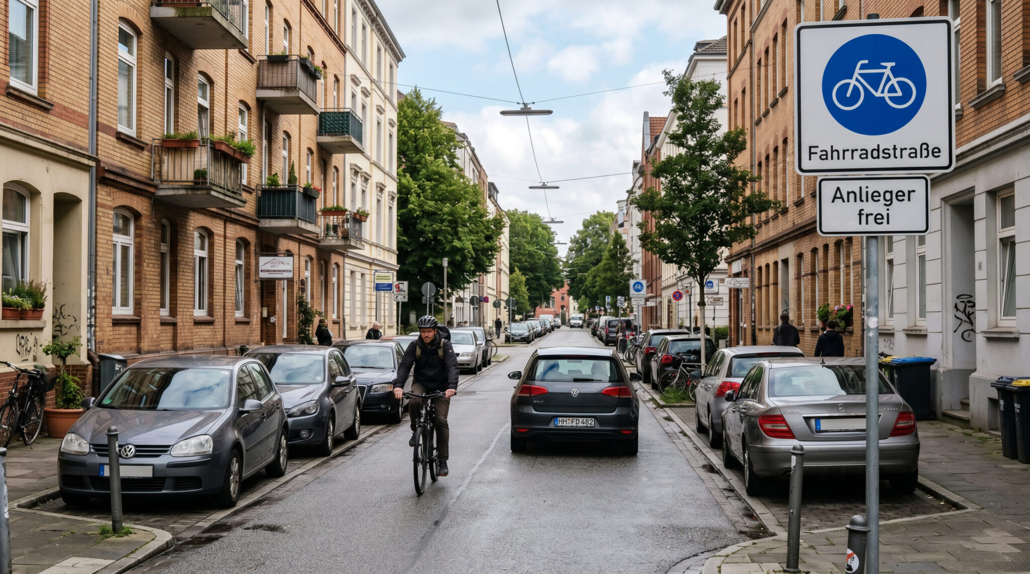 Blaues Fahrradstraßen-Schild in einer engen Wohnstraße mit parkenden Autos, einem Radfahrer und einem PKW dahinter.
